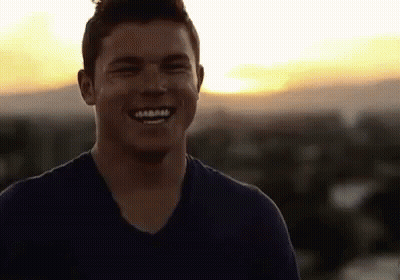 A Man In A Blue Shirt Is Smiling At The Camera While Standing On Top Of A Building At Sunset . GIF