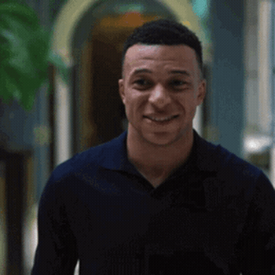 A Man In A Blue Shirt Is Smiling While Standing In A Hallway . GIF