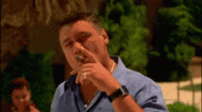 A Man In A Blue Shirt Is Smoking A Cigar Outdoors GIF