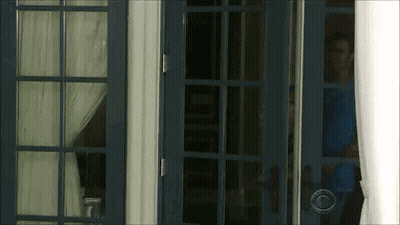 A Man In A Blue Shirt Is Standing In A Doorway Looking Out . GIF