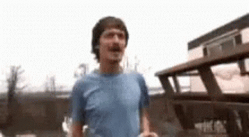 A Man In A Blue Shirt Is Standing In Front Of A Broken Down Bus . GIF