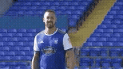A Man In A Blue Shirt Is Standing In Front Of A Stadium . GIF