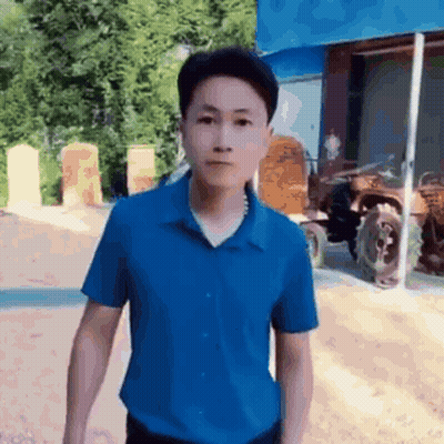 A Man In A Blue Shirt Is Standing In Front Of A Tractor And Looking At The Camera . GIF