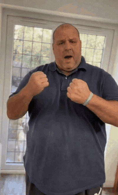 A Man In A Blue Shirt Is Standing In Front Of A Window With His Fists Up . GIF
