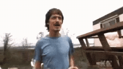 A Man In A Blue Shirt Is Standing In Front Of A Wooden Building . GIF