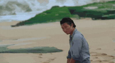 A Man In A Blue Shirt Is Standing On A Sandy Beach Looking At Something GIF