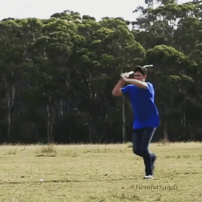 A Man In A Blue Shirt Is Swinging A Golf Club In A Field With Trees In The Background GIF