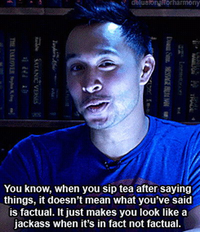 A Man In A Blue Shirt Is Talking In Front Of A Shelf Of Books GIF