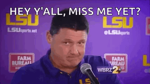 A Man In A Blue Shirt Is Talking Into A Microphone In Front Of A Purple Wall . GIF