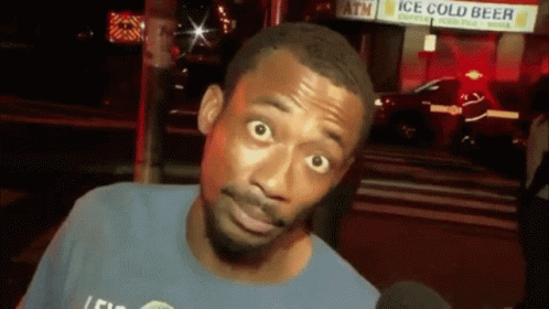 A Man In A Blue Shirt Is Talking Into A Microphone In Front Of A Sign That Says Ice Cold Beer GIF