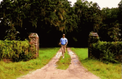 A Man In A Blue Shirt Is Walking Down A Dirt Road . GIF