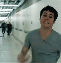 A Man In A Blue Shirt Is Walking Down A Hallway With People Walking In The Background . GIF