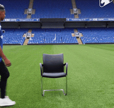 A Man In A Blue Shirt Is Walking On A Soccer Field With A Chair In The Foreground That Says Kbb Centre GIF