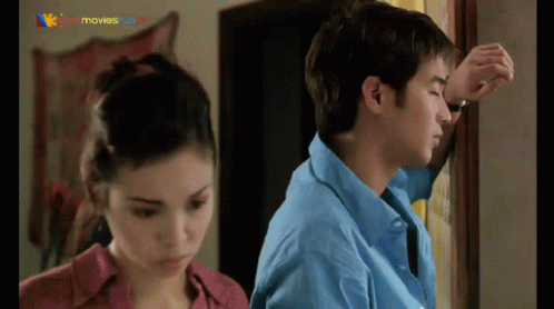 A Man In A Blue Shirt Leans Against A Wall While A Woman In A Red Shirt Looks On GIF