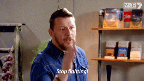A Man In A Blue Shirt Says Stop Fighting In Front Of Some Shelves GIF