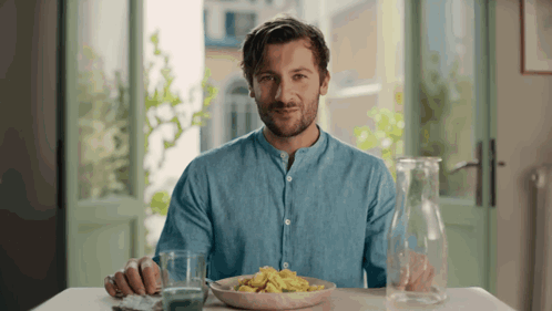 A Man In A Blue Shirt Sits At A Table With A Plate Of Food GIF