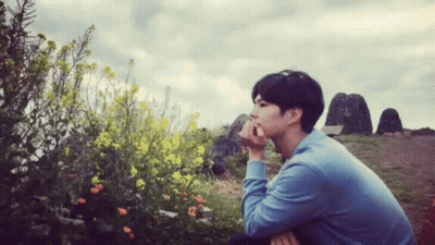A Man In A Blue Shirt Sits In Front Of Flowers GIF