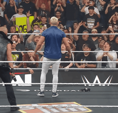 A Man In A Blue Shirt Stands In A Wrestling Ring With A Crowd Behind Him GIF