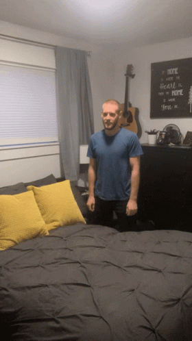 A Man In A Blue Shirt Stands In Front Of A Bed With A Sign That Says Home GIF