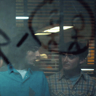 A Man In A Blue Shirt Stands Next To Another Man In A Plaid Shirt In Front Of A Window GIF