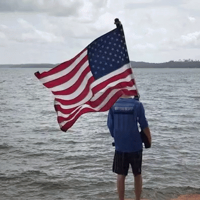 A Man In A Blue Shirt That Says Watercraft Holds An American Flag GIF