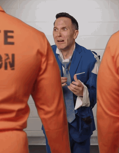 A Man In A Blue Suit And Tie Is Standing Next To A Man In An Orange Uniform With The Letter E On The Back GIF