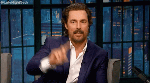 A Man In A Blue Suit And White Shirt Is Sitting In A Chair And Pointing At The Camera . GIF