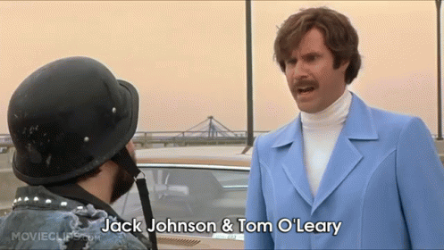 A Man In A Blue Suit And White Turtleneck Is Talking To Another Man In A Black Helmet .. GIF
