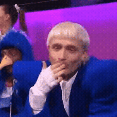 A Man In A Blue Suit Is Covering His Mouth With His Hand While Sitting In Front Of A Purple Wall . GIF