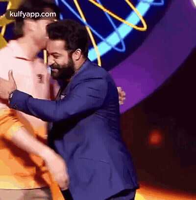 A Man In A Blue Suit Is Hugging Another Man In An Orange Shirt On A Stage . GIF