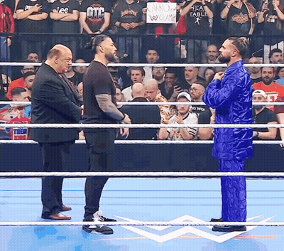 A Man In A Blue Suit Stands In The Middle Of A Wrestling Ring With Two Other Men GIF