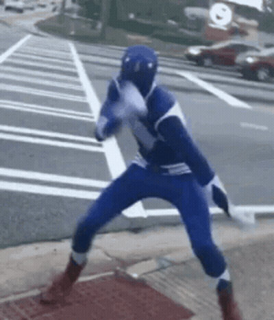 A Man In A Blue Superhero Costume Is Standing On The Sidewalk . GIF