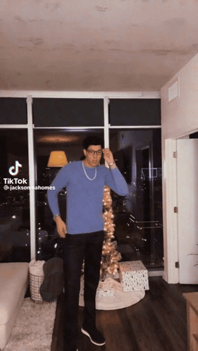 A Man In A Blue Sweater And Black Pants Is Standing In Front Of A Christmas Tree In A Living Room GIF