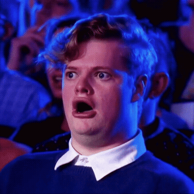 A Man In A Blue Sweater And White Collar Is Making A Surprised Face While Sitting In A Crowd . GIF