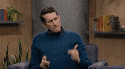 A Man In A Blue Sweater Is Sitting In A Chair Making A Gesture With His Hands . GIF