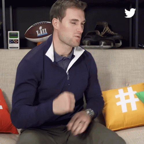 A Man In A Blue Sweater Is Sitting On A Couch With A Pillow That Says # On It GIF