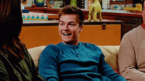 A Man In A Blue Sweater Is Smiling While Sitting On A Couch . GIF