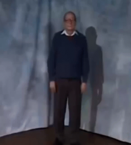 A Man In A Blue Sweater Is Standing In Front Of A Blue Curtain . GIF
