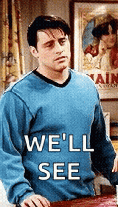 A Man In A Blue Sweater Is Standing Next To A Table With The Words `` We 'Ll See '' Written On It . GIF