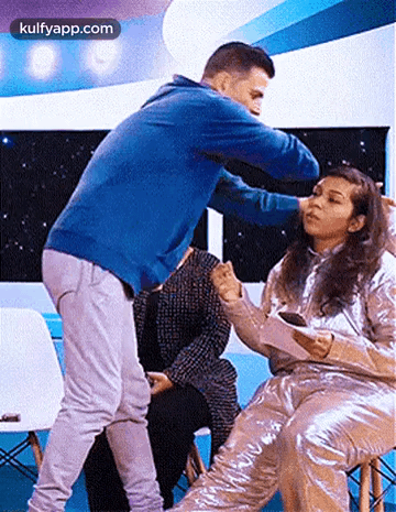 A Man In A Blue Sweater Is Standing Next To A Woman In A Silver Outfit . GIF