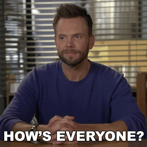 A Man In A Blue Sweater Sits At A Table With The Words How 'S Everyone Written Below Him GIF