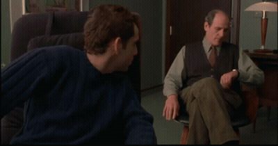A Man In A Blue Sweater Sits In A Chair Next To Another Man In A Vest And Tie GIF