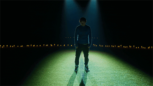 A Man In A Blue Sweater Stands In A Dark Room GIF