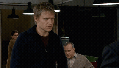 A Man In A Blue Sweater Stands In A Dark Room With Other Men GIF