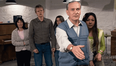 A Man In A Blue Vest Stands In Front Of A Group Of People Including A Woman Playing A Guitar GIF