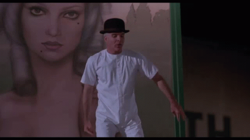 A Man In A Bowler Hat Is Standing In Front Of A Sign That Says Look Up Here GIF