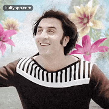 A Man In A Brown And White Striped Sweater Is Smiling In Front Of Flowers . GIF