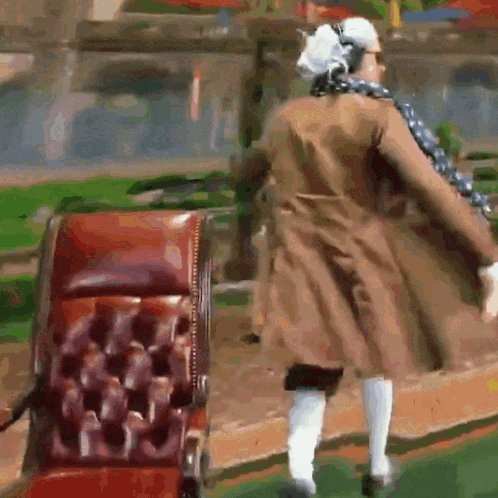 A Man In A Brown Coat And White Socks Is Walking In Front Of A Red Chair GIF
