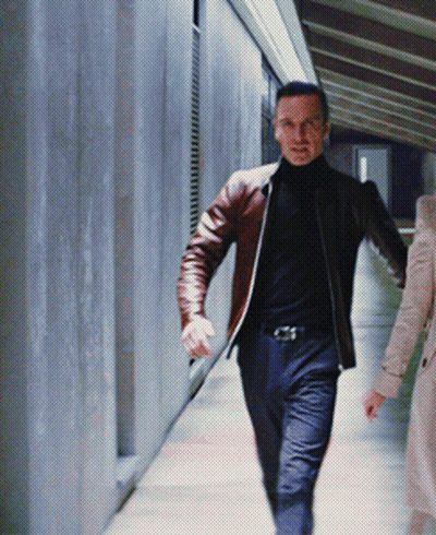 A Man In A Brown Jacket And Black Turtleneck Is Walking Down A Hallway GIF