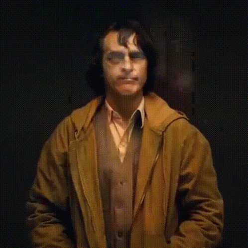 A Man In A Brown Jacket And Orange Vest Is Making A Funny Face While Standing In A Dark Room . GIF